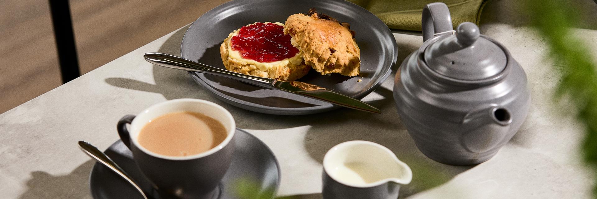 Grey teapot, cup of Breakfast Tea with milk, and a fruit scone with jam and cream on a white marble table.